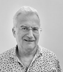 Portrait d'Urb1n, homme âgé souriant portant des lunettes et une chemise à motifs.